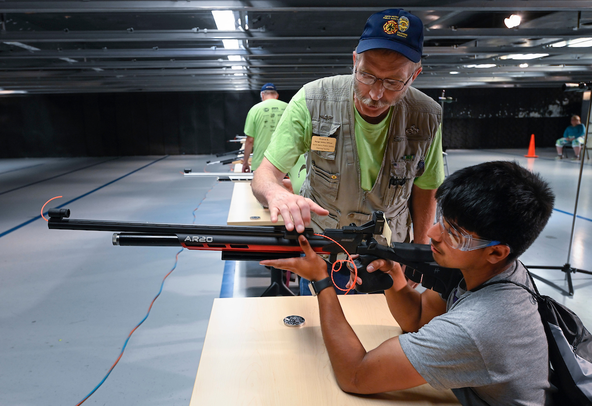 A young adaptive athlete is aiming a rifle and being coached by an older person standing behind them