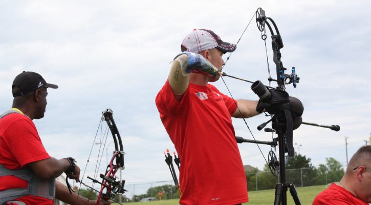 A person with a right arm amputation participating in outdoor archery, drawing back a bow and aiming at a target.