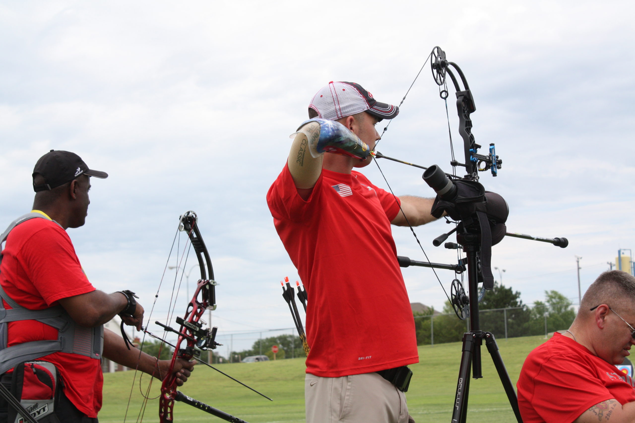 three athletes wearing red shirts competing in archery. One athlete uses a prosthetic arm; an athlete using a wheelchair aims his bow while an onlooker watches; a woman using a wheelchair aims her bow at a target in the middle of a grassy field; an adaptive athlete using his shoulder and foot to hold and aim his bow during an archery competition