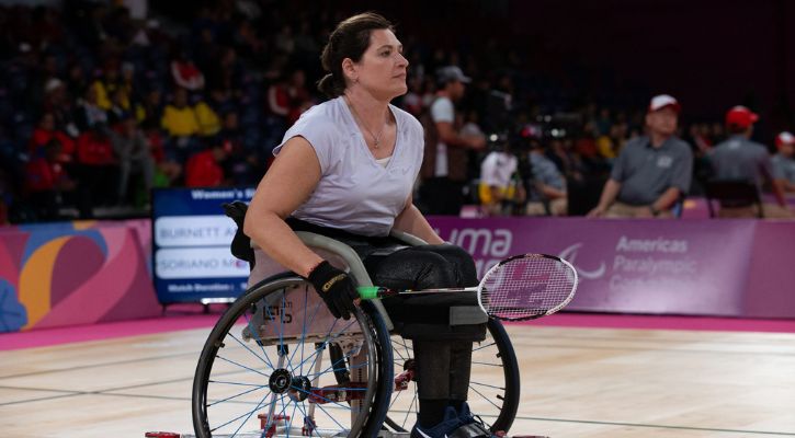 A woman in a wheelchair at an indoor court participating in a game of badmitton