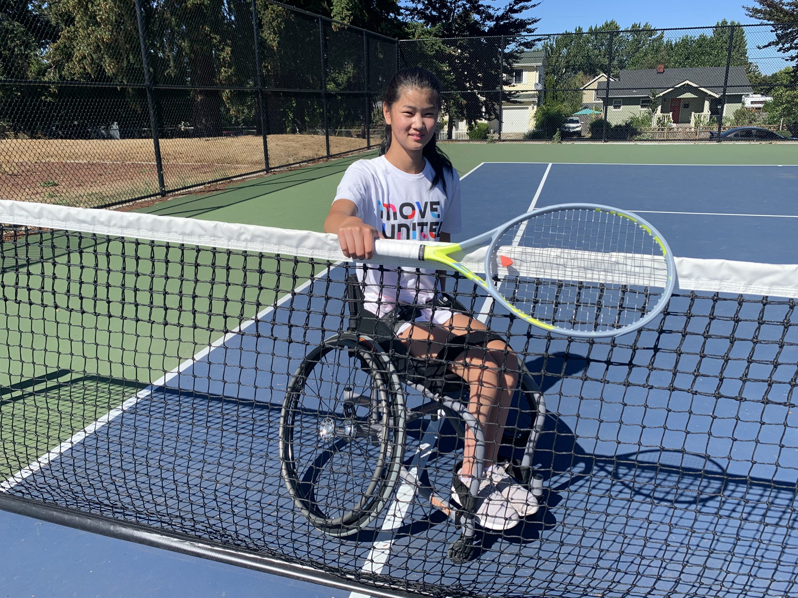 Female athlete in a wheelchair behind a tennis net holding a tennis racket