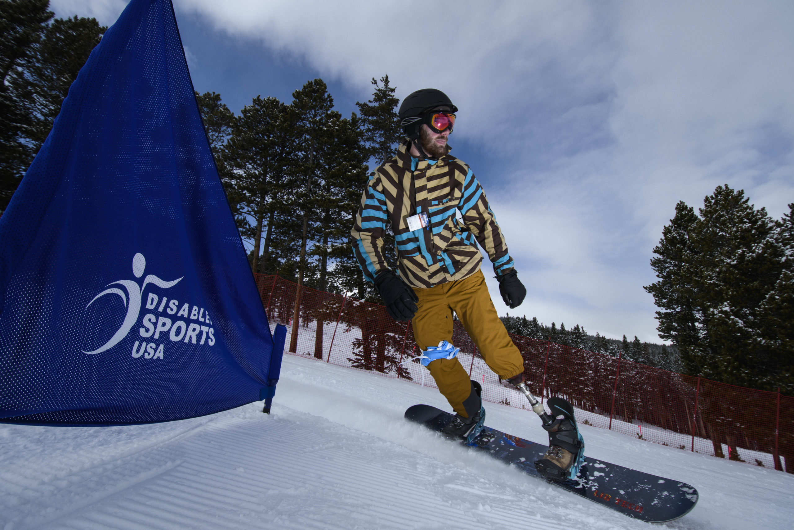 Male athlete with a left leg below the knee amputation skiing down hill past a banner