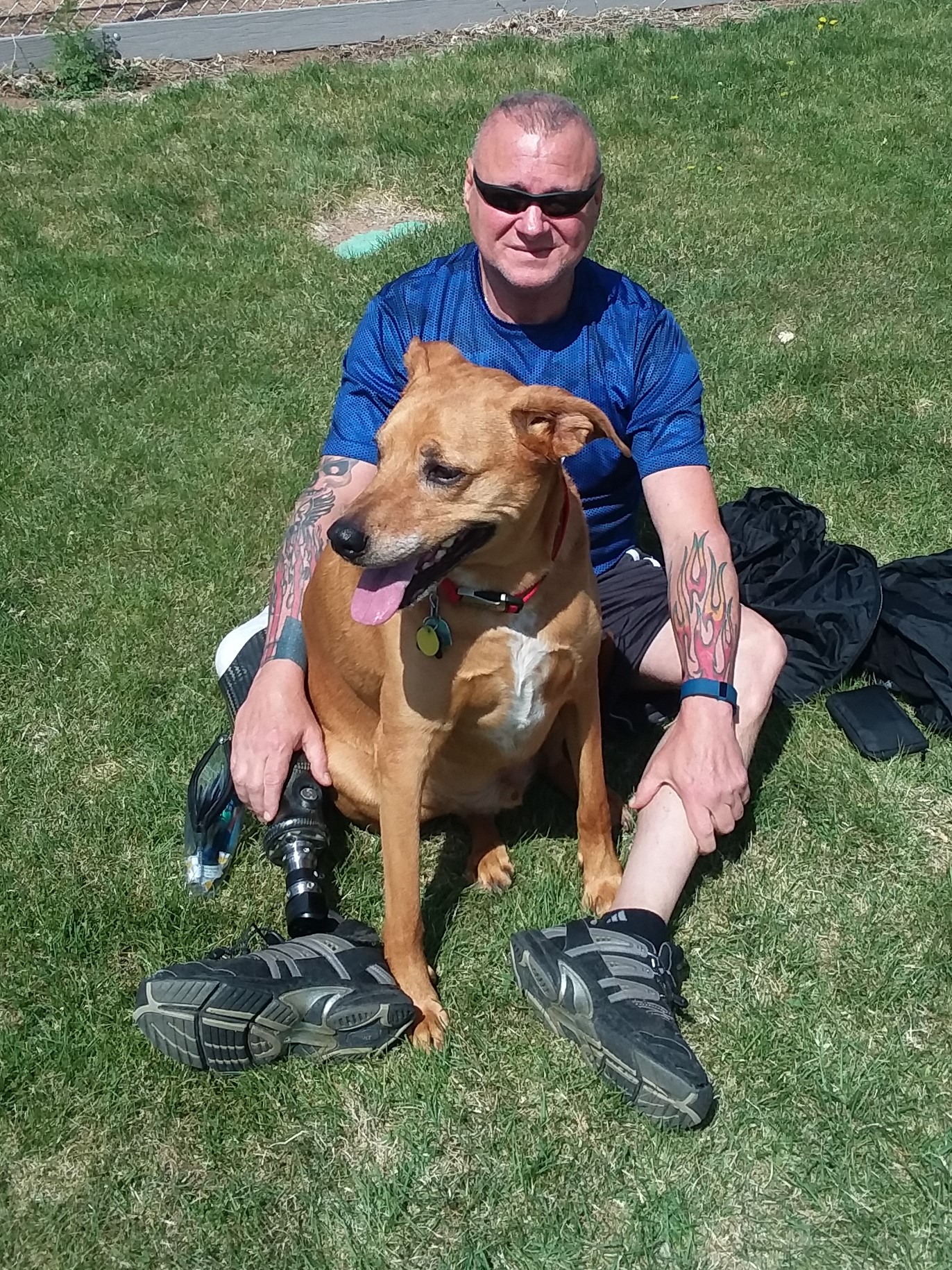 Male sitting on the ground with right leg prosthetic smiling at the camera with dog sitting in between his legs
