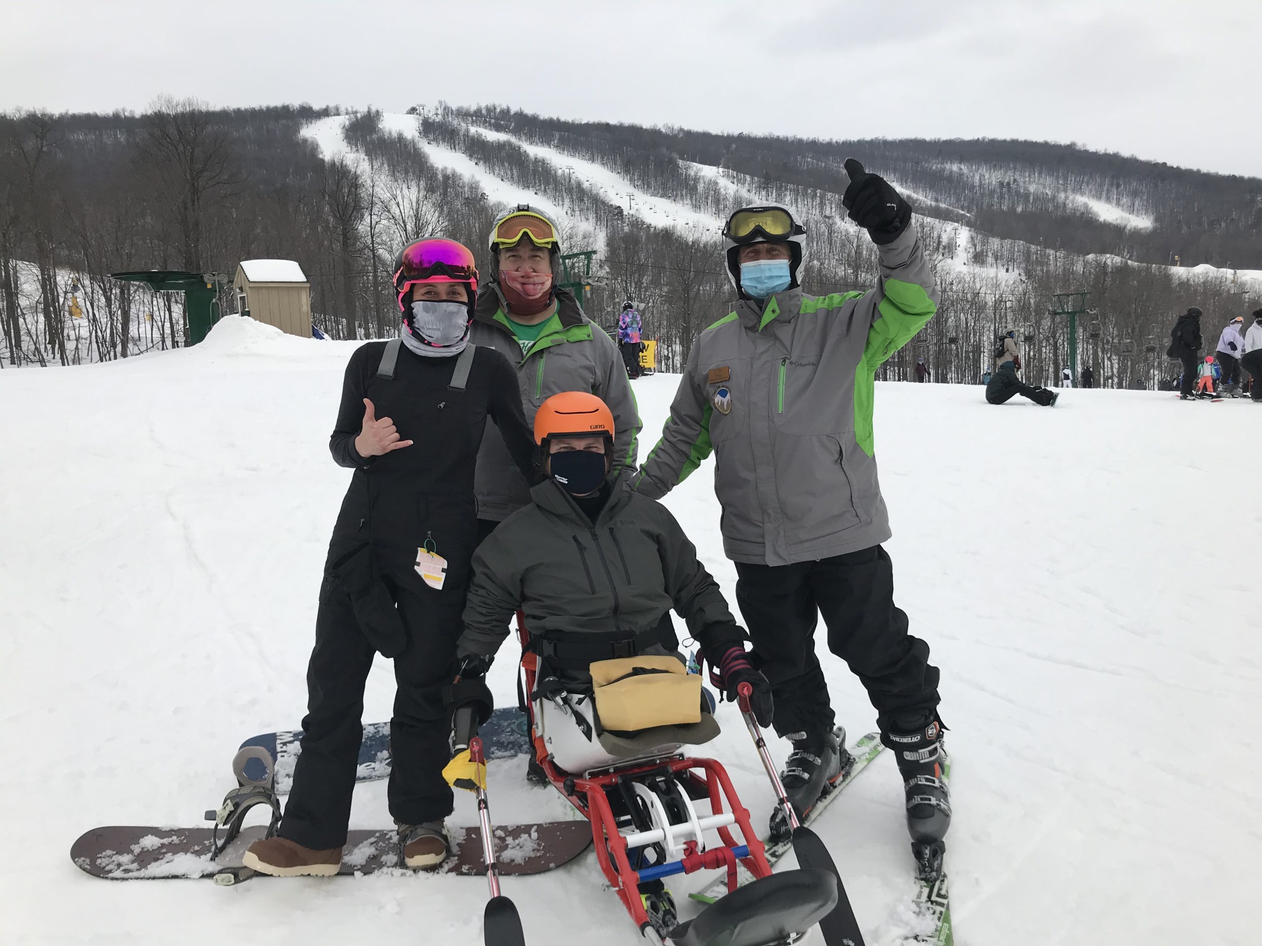 Group of athletes with snowboards, skis, and mono-skis smiling at the camera and giving a thumbs up