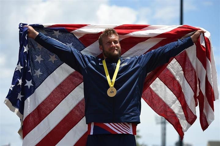 Male athlete wearing a medal smiling and holding an American flag behind him