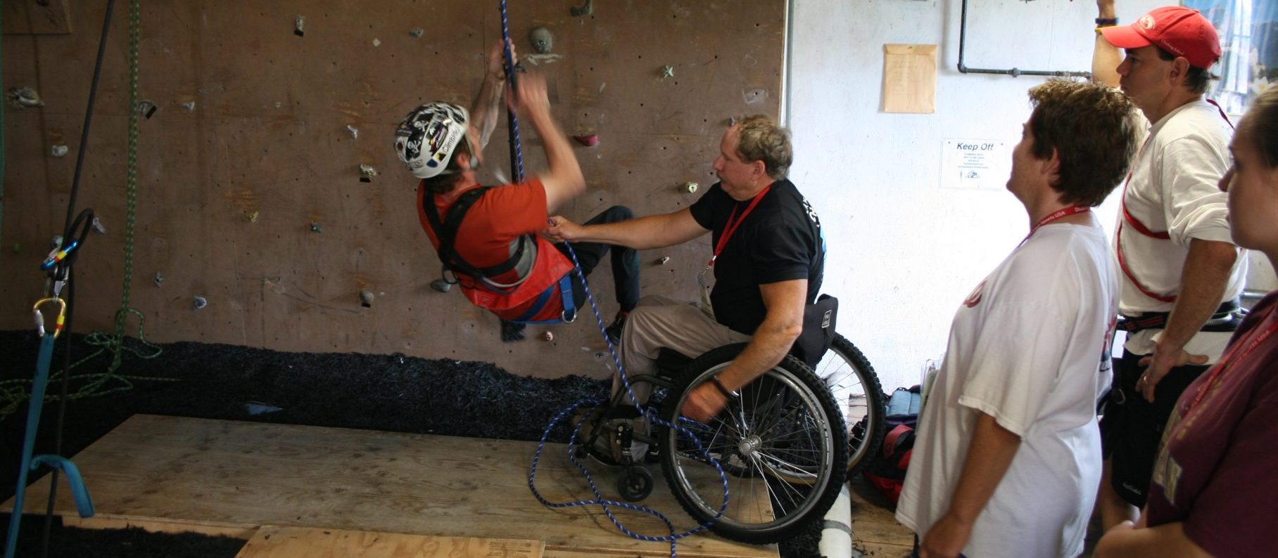 Athlete climbing on rock wall with assistance of instructor in a wheelchair with other participants onlooking