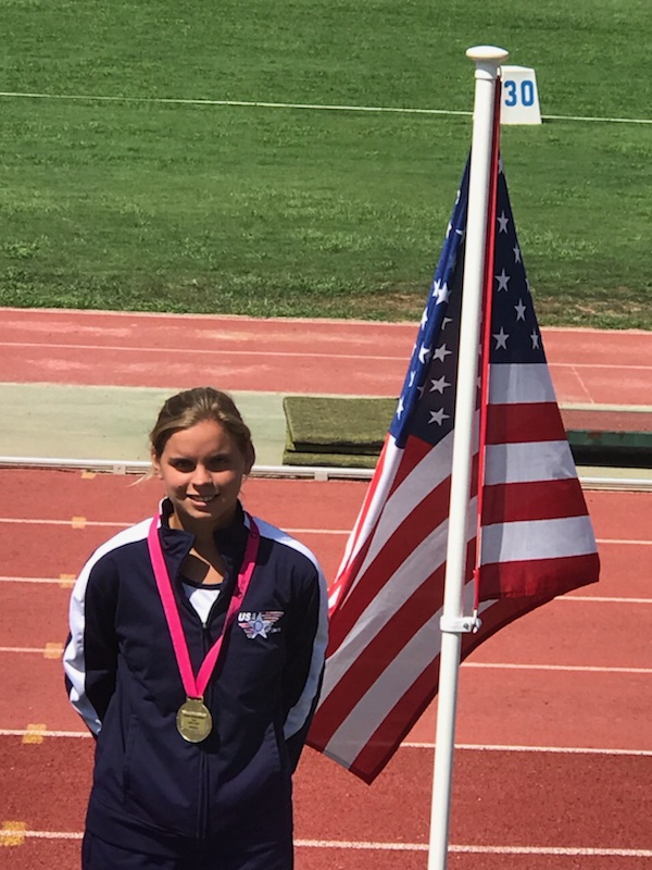 Female athlete with a medal posed smiling at the camera standing on a running track next to an American flag