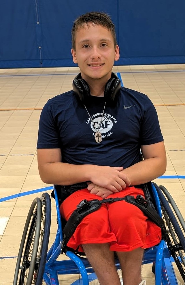 A young man in a wheelchair smiles at the camera in a gym. He is wearing a navy blue T-shirt with the Challenged Athletes Foundation logo, bright red shorts, and headphones resting around his neck.