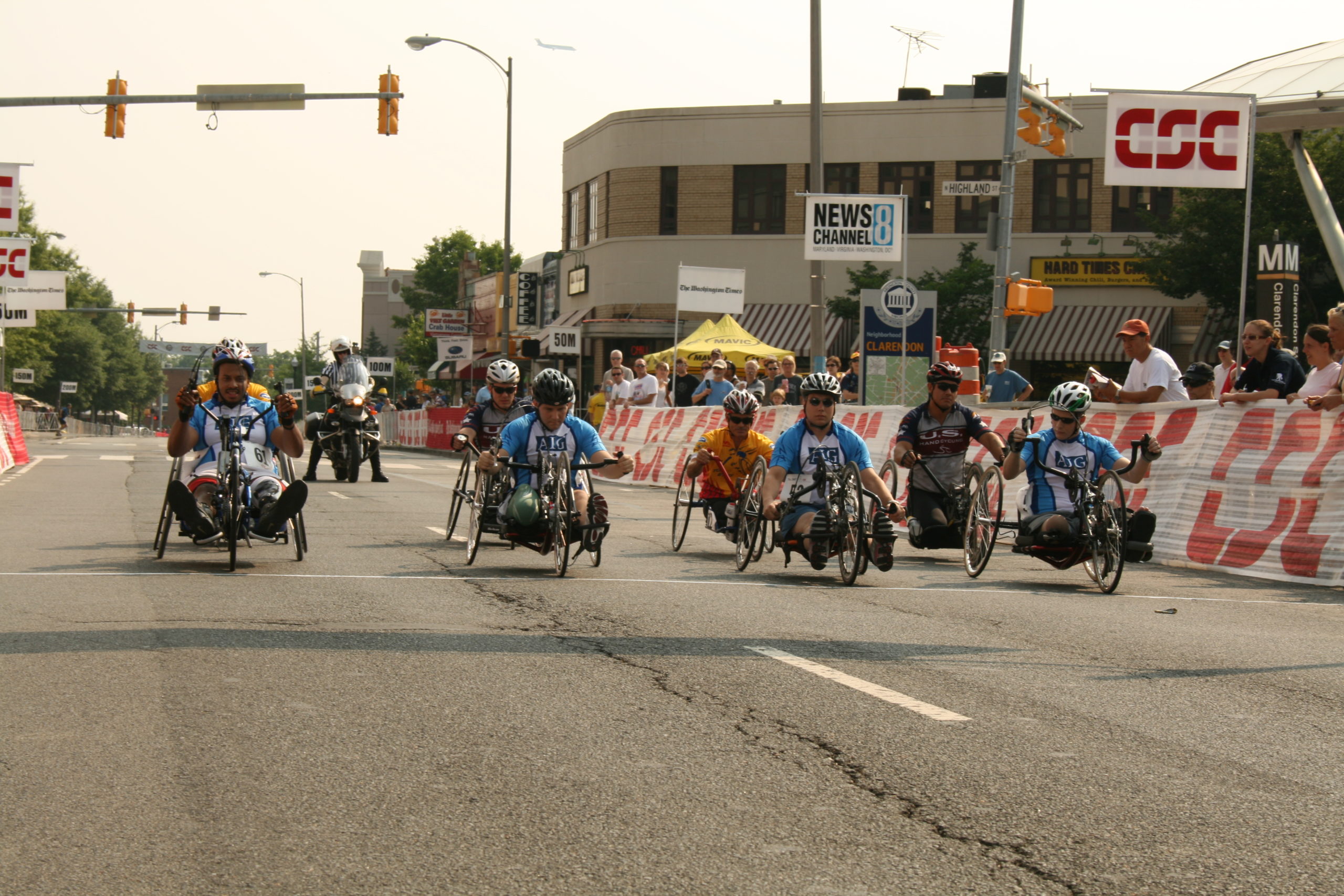 Group of athletes competing in hand cycling against each other on a street with onlookers to their left