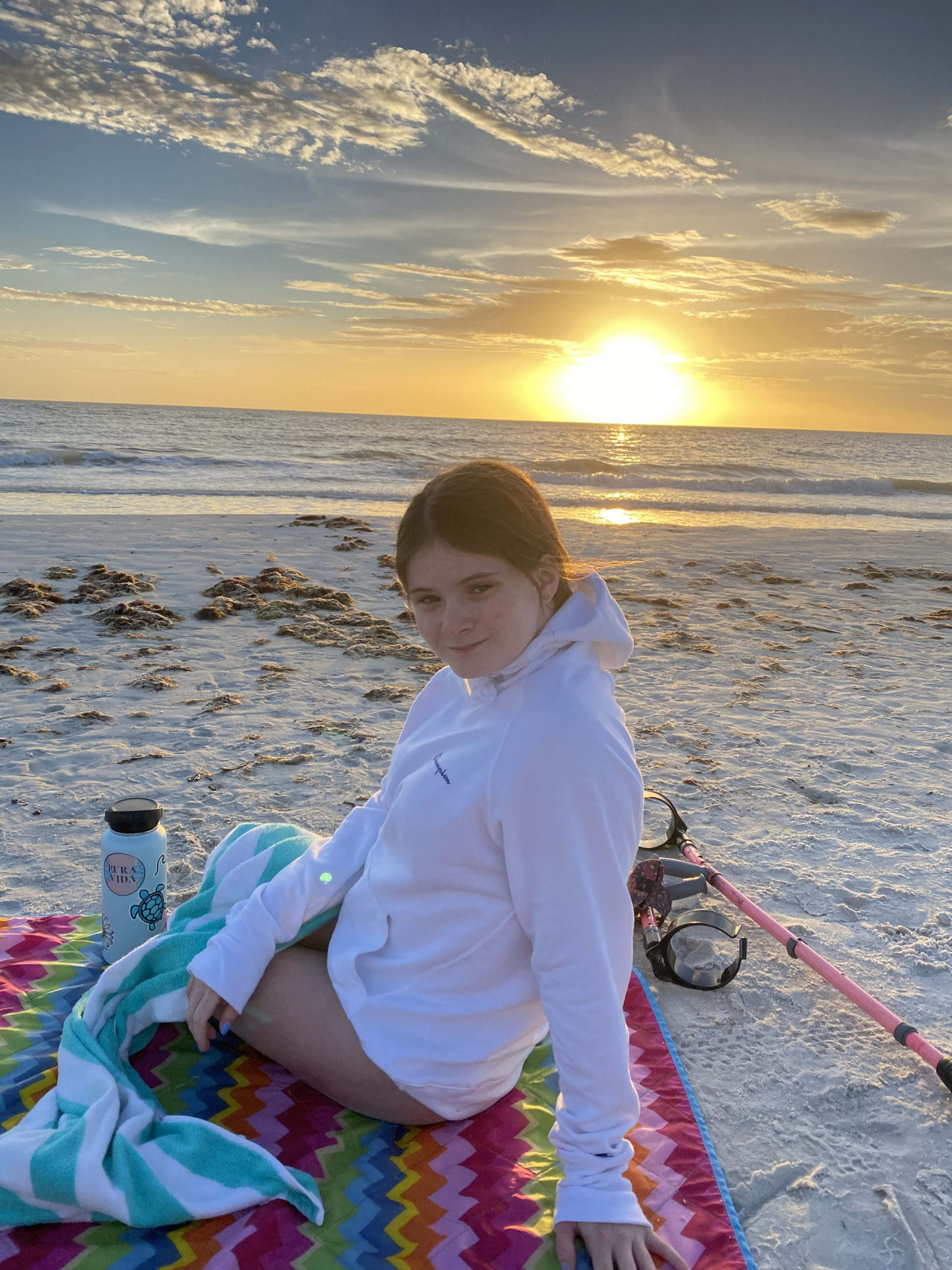 Female sitting on a beach towel at the beach turned around smiling at the camera with sunrise in the background