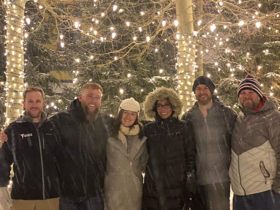 Group of people smiling at the camera with snow and lights wrapped around trees in the background