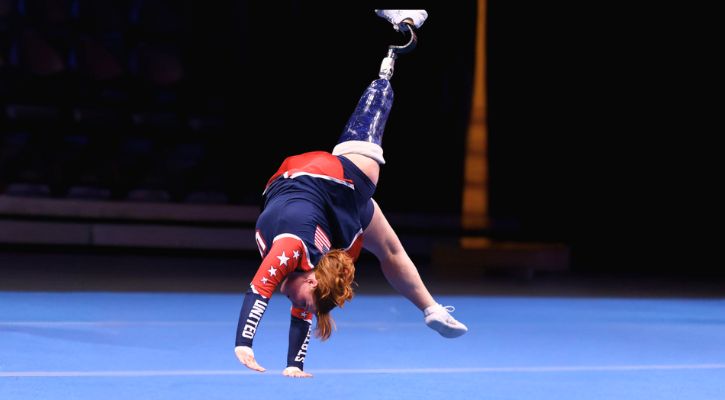 A gymnast with a prosthetic leg performing a cartwheel on a blue floor.