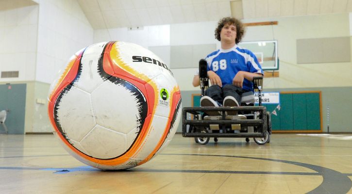 A power soccer player in a blue jersey maneuvers their wheelchair behind a large soccer ball on an indoor court.