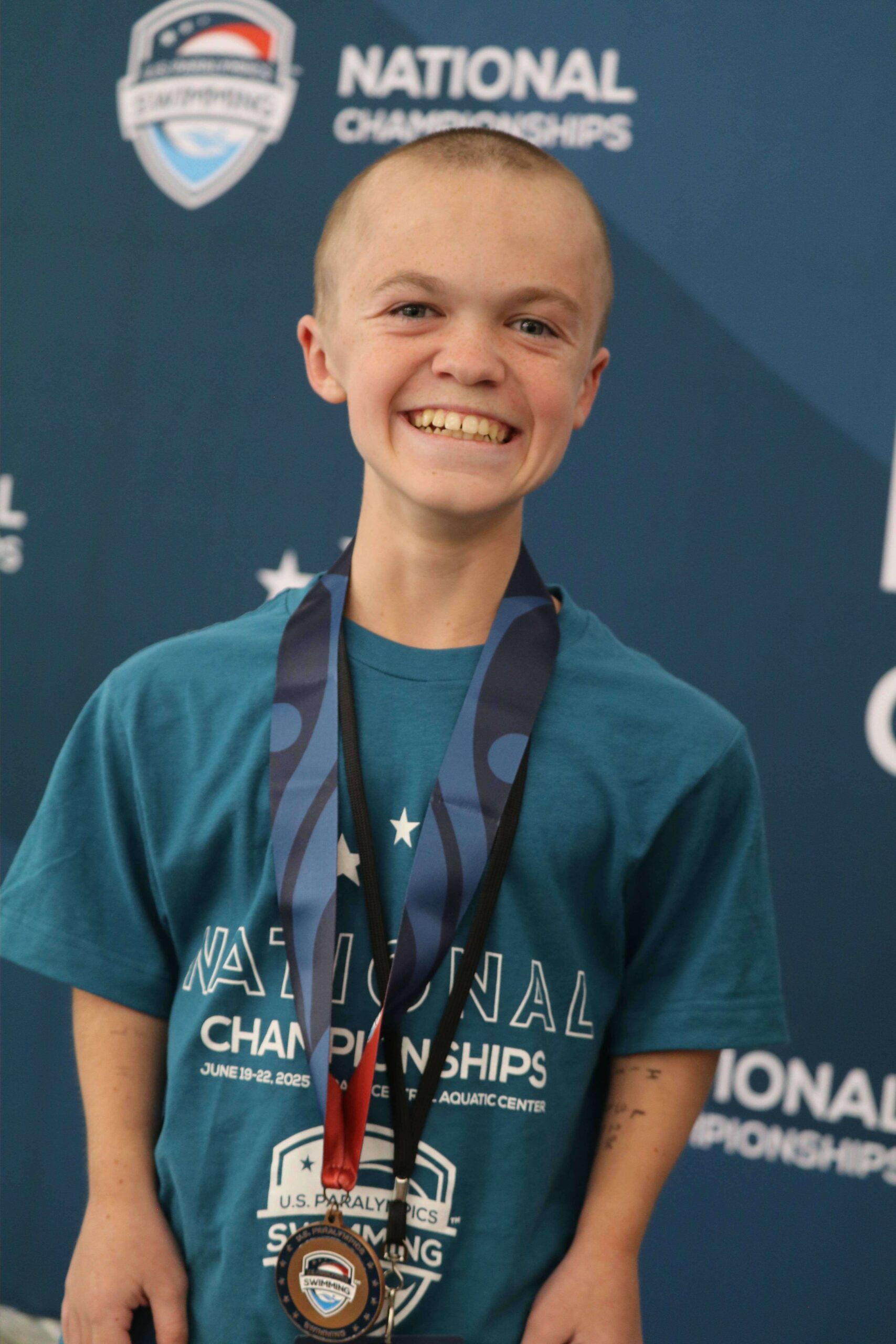A young adaptive athlete wearing a teal “National Championships” T-shirt smiles proudly while displaying his medals at the 2025 U.S. Paralympics Swimming National Championships. The event logo and backdrop are visible behind him.