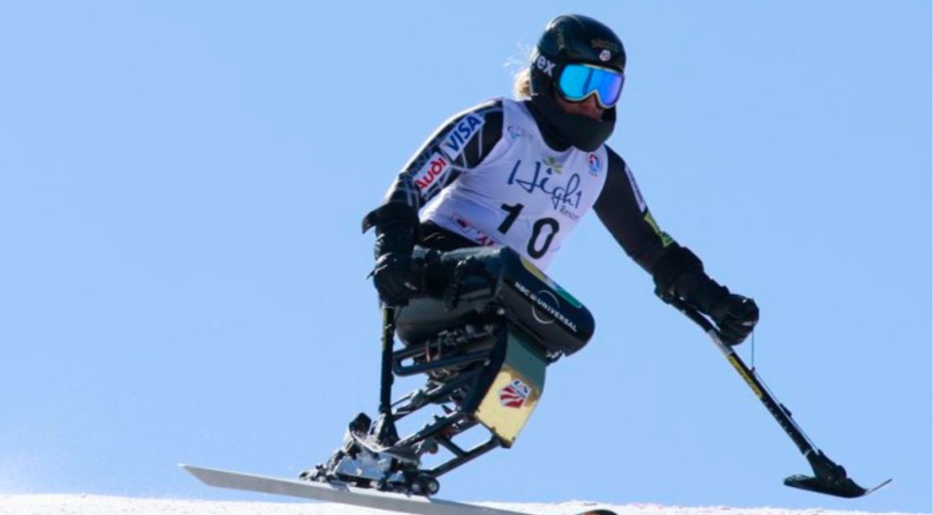 A Move United adaptive skier competes in a para alpine skiing event, racing downhill in a sit-ski under a clear blue sky while wearing a black helmet, reflective goggles, and a white race bib with the number 10.