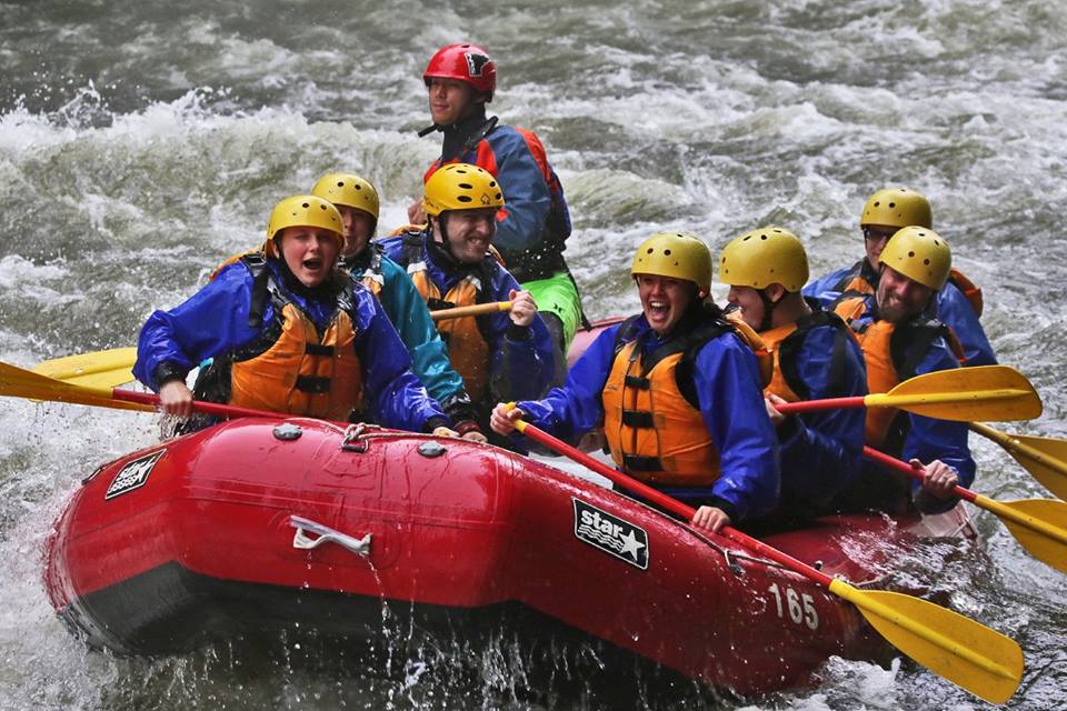 Group of athletes smiling and rowing on a boat through white water rapids