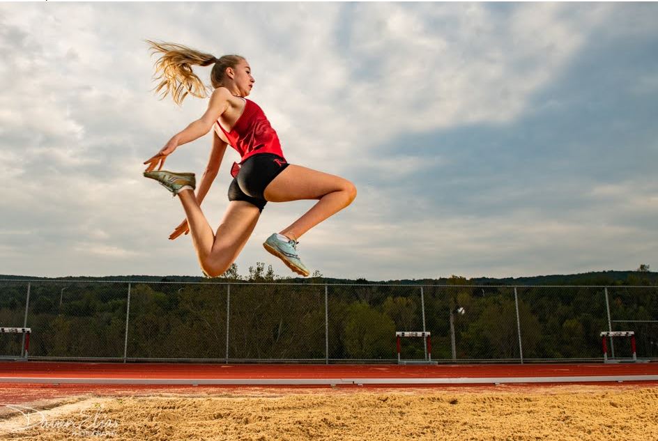 White female mid long jump with long blonde ponytail behind her with a red top and black shorts