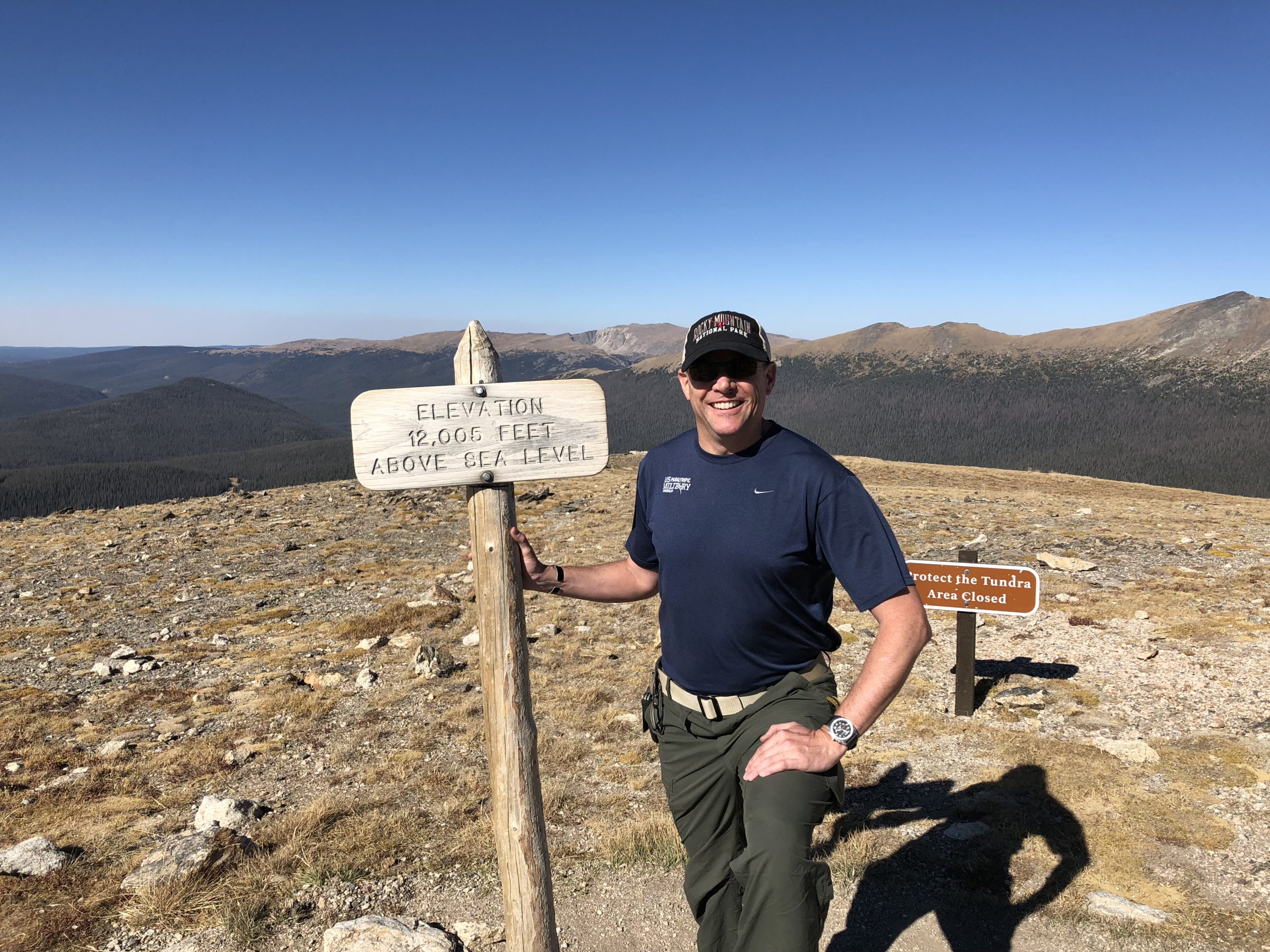 Male posed smiling at the camera by a sign that says elevation 12,000 feet above sea level with mountains in the background