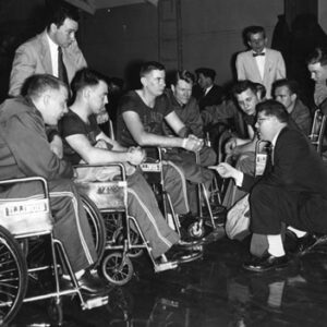 black and white photo of a team of male athletes in wheelchairs listening to their coach who is kneeling on the ground