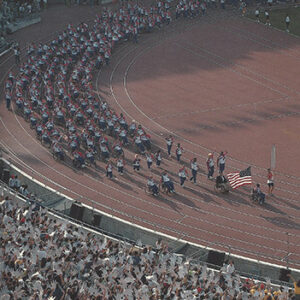 Athletes in wheelchairs around a track lead by someone with an American flag