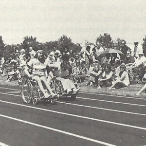black and white photo of two athletes in wheelchairs racing on a track with a crowd of people watching