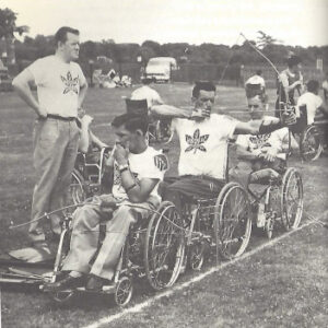 black and white photo of three athletes in wheelchairs doing archery with a coach standing behind them