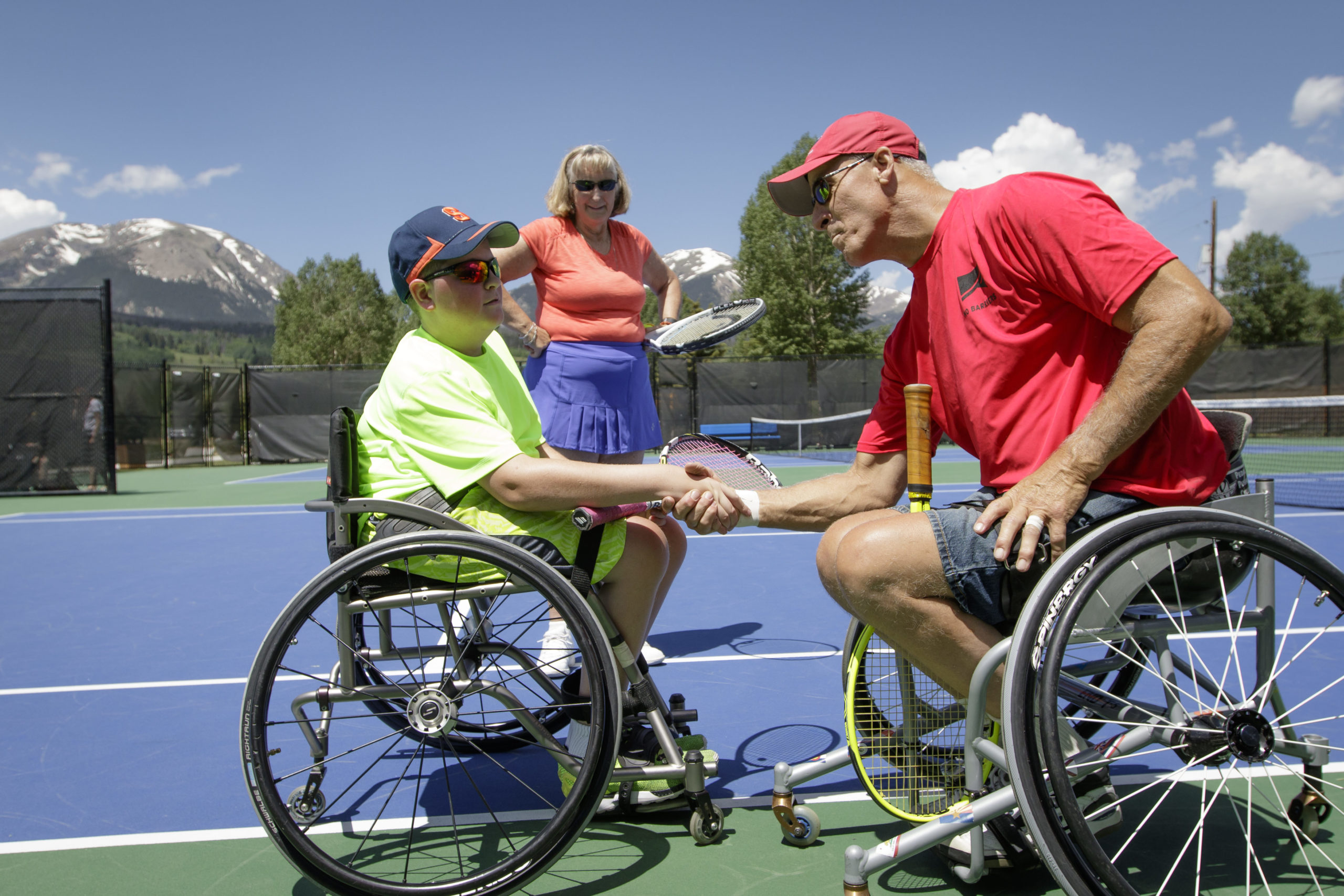 Two athletes in wheelchairs shaking hands with woman standing behind them watching