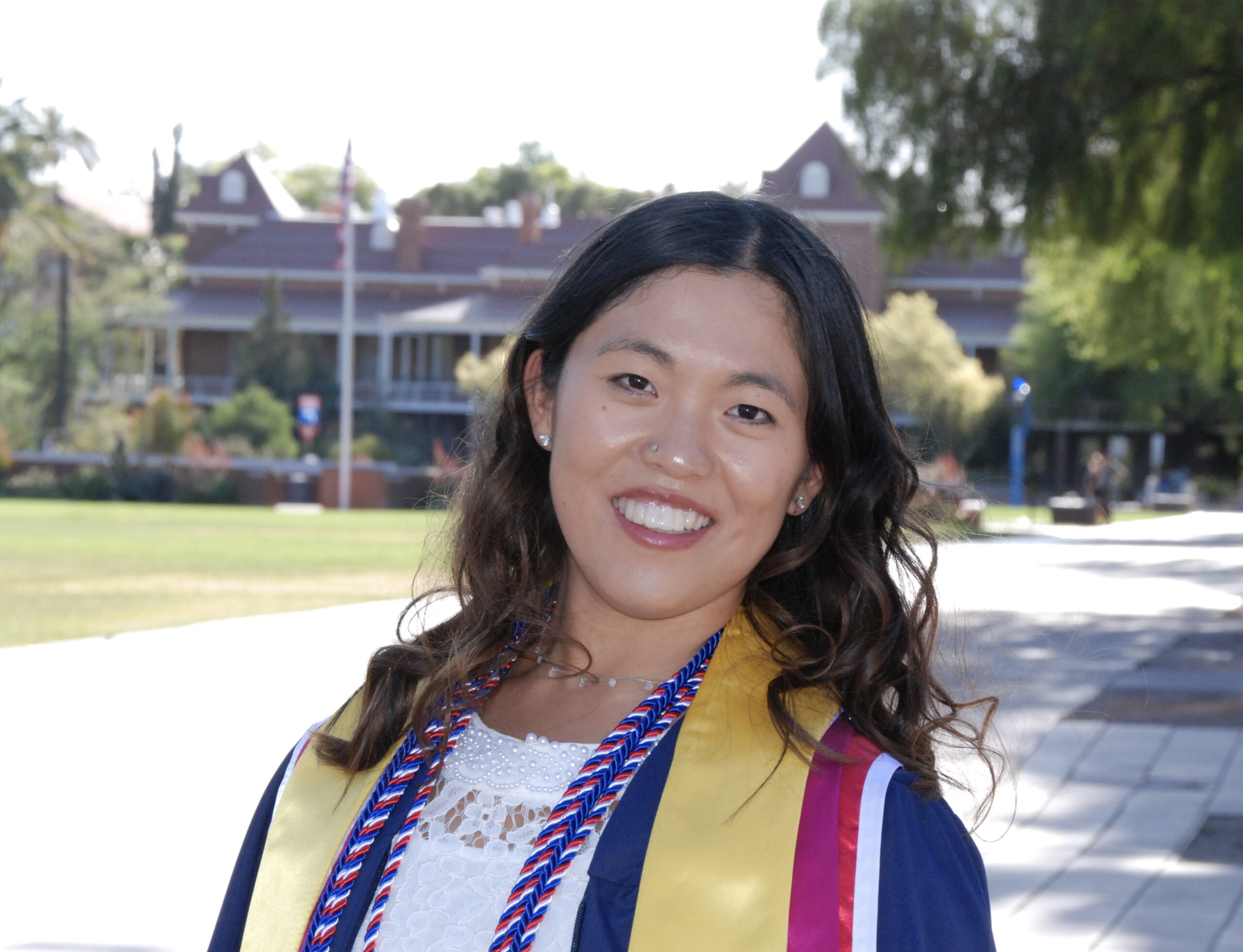 Move United Member Athlete Jacie King smiles outdoors in her graduation attire, wearing a navy gown with honor cords and a yellow stole. Trees, a walkway, and a campus building appear in the background on a sunny day.
