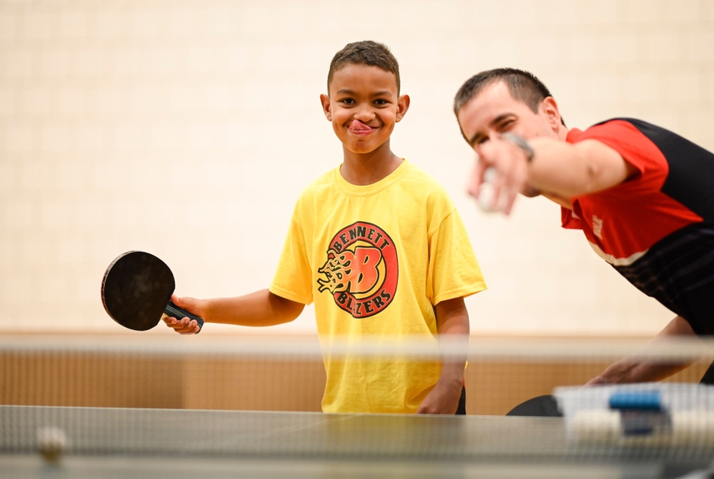 Young male athlete holding table tennis racket smiling with his tongue out at the camera with his coach beside him pointing to the camera