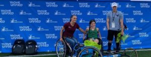 Three athletes in front of a step and repeat in wheelchairs with one athlete standing holding a bow and arrow and a racing wheelchair in front of the athletes