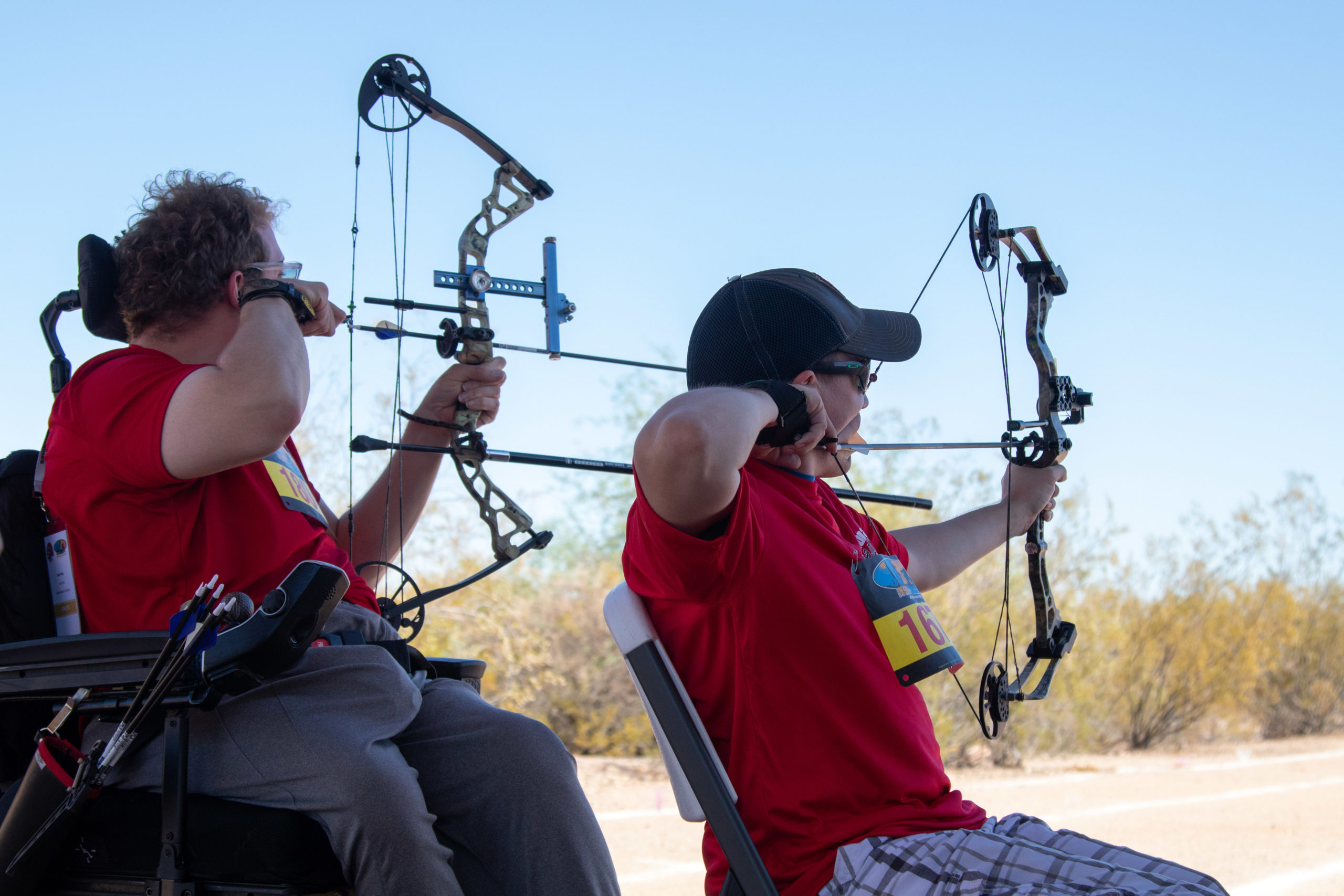 Two athletes one in a wheelchair and the other sitting aiming a bow and arrow