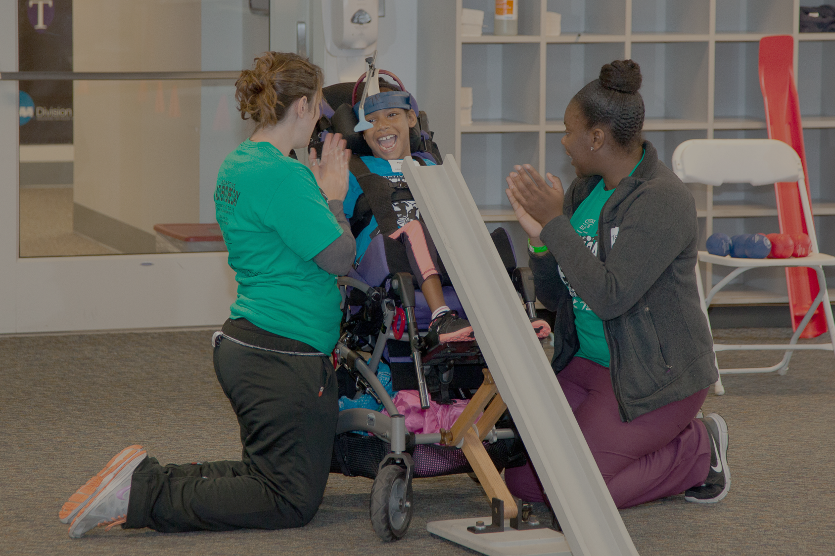 Child in wheelchair playing boccia with two other people assisting and giving praise