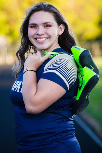 Female athlete wearing a blue shirt smiling at the camera holding green athletic shoes
