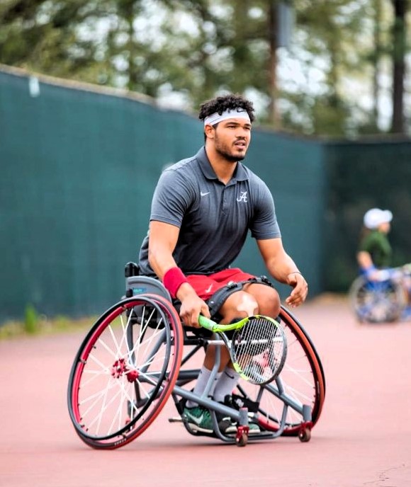 Move United member athlete Jeremy Boyd plays wheelchair tennis on an outdoor court, maneuvering his sport wheelchair while holding a tennis racquet. He wears a gray polo, red shorts, and a headband, with a green fence and trees in the background.