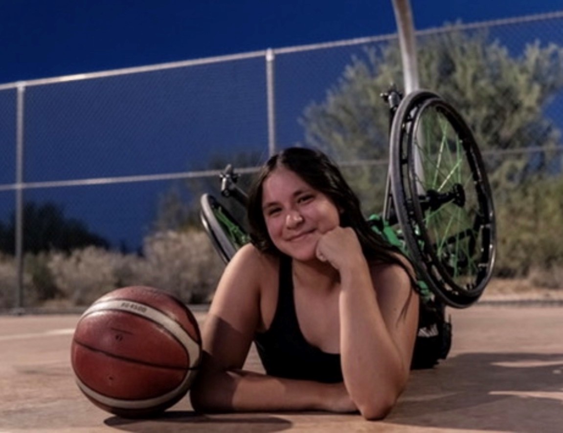 Adrina Castro, a Move United athlete, smiles while lying on a basketball court beside her wheelchair and a basketball. The photo is taken outdoors at dusk, with a chain-link fence and trees in the background.