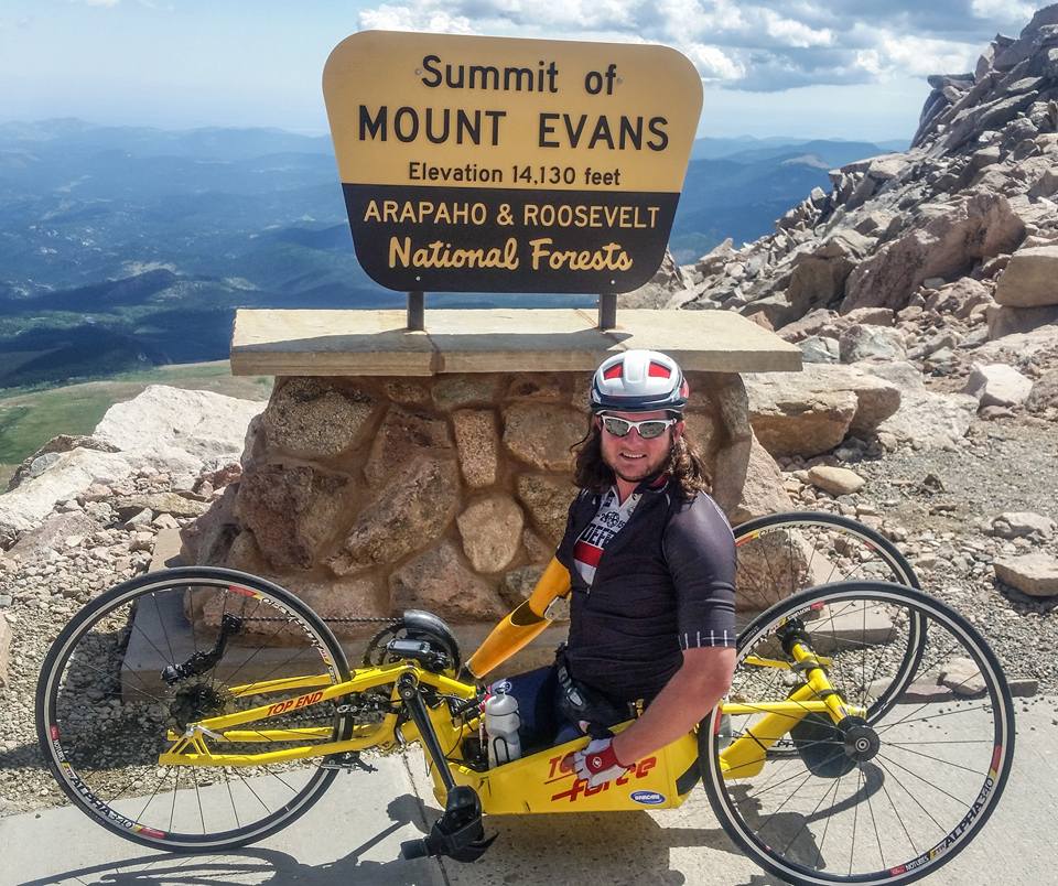 Tim Brown poses in front of the summit sign for Mount Evans. He is wearing black cycling kit and sitting in a yellow kneeler handcycle.