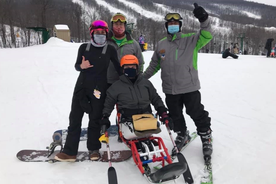 Tim Brown, wearing a green ski jacket and orange helmet, poses with Jamie Rihn and instructors from Two Top Mountain on a ski run. Tim is sitting in a Monique monoski.