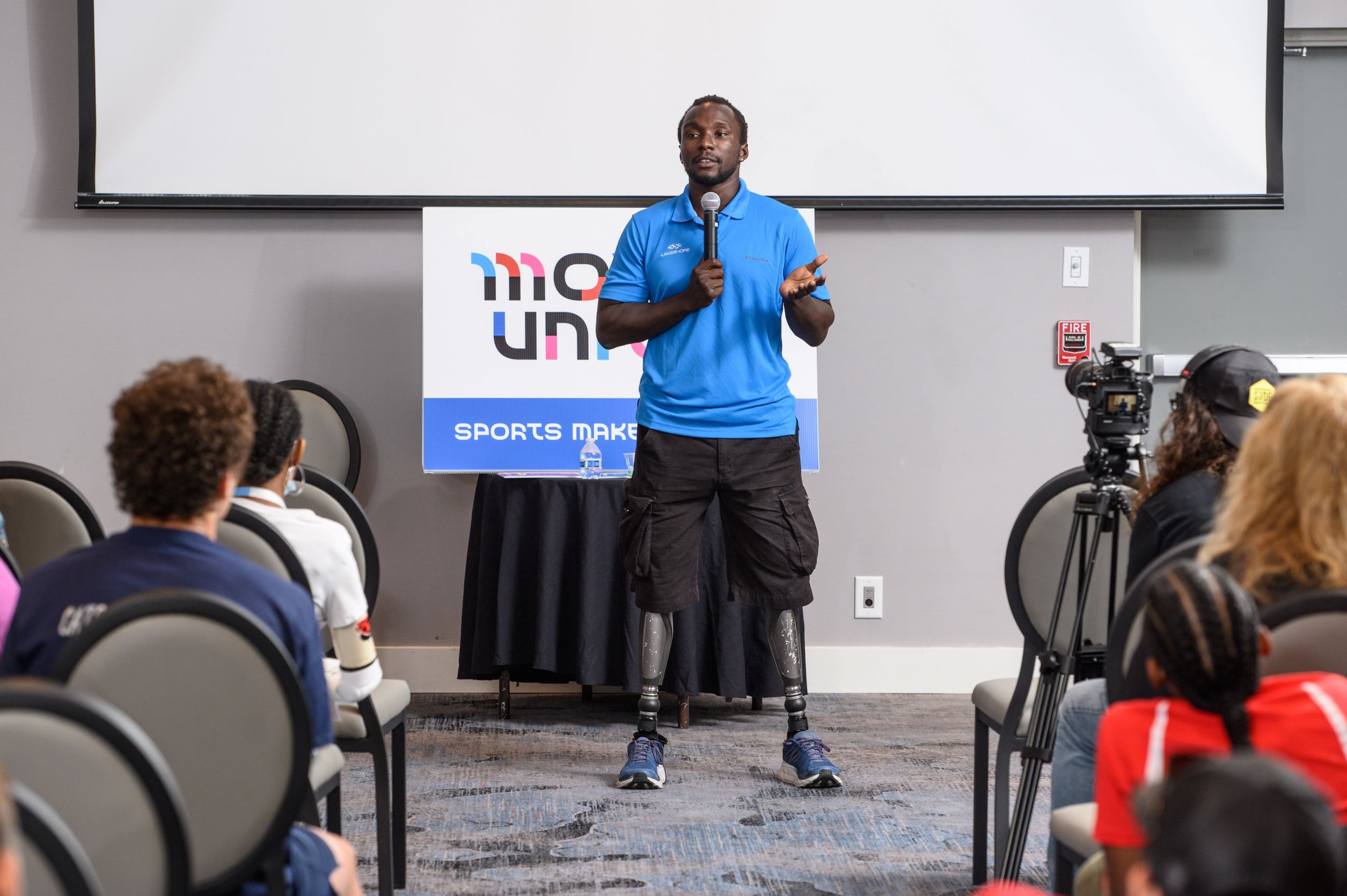 Presenter with double leg amputation speaking in front of audience with Move United logo behind him