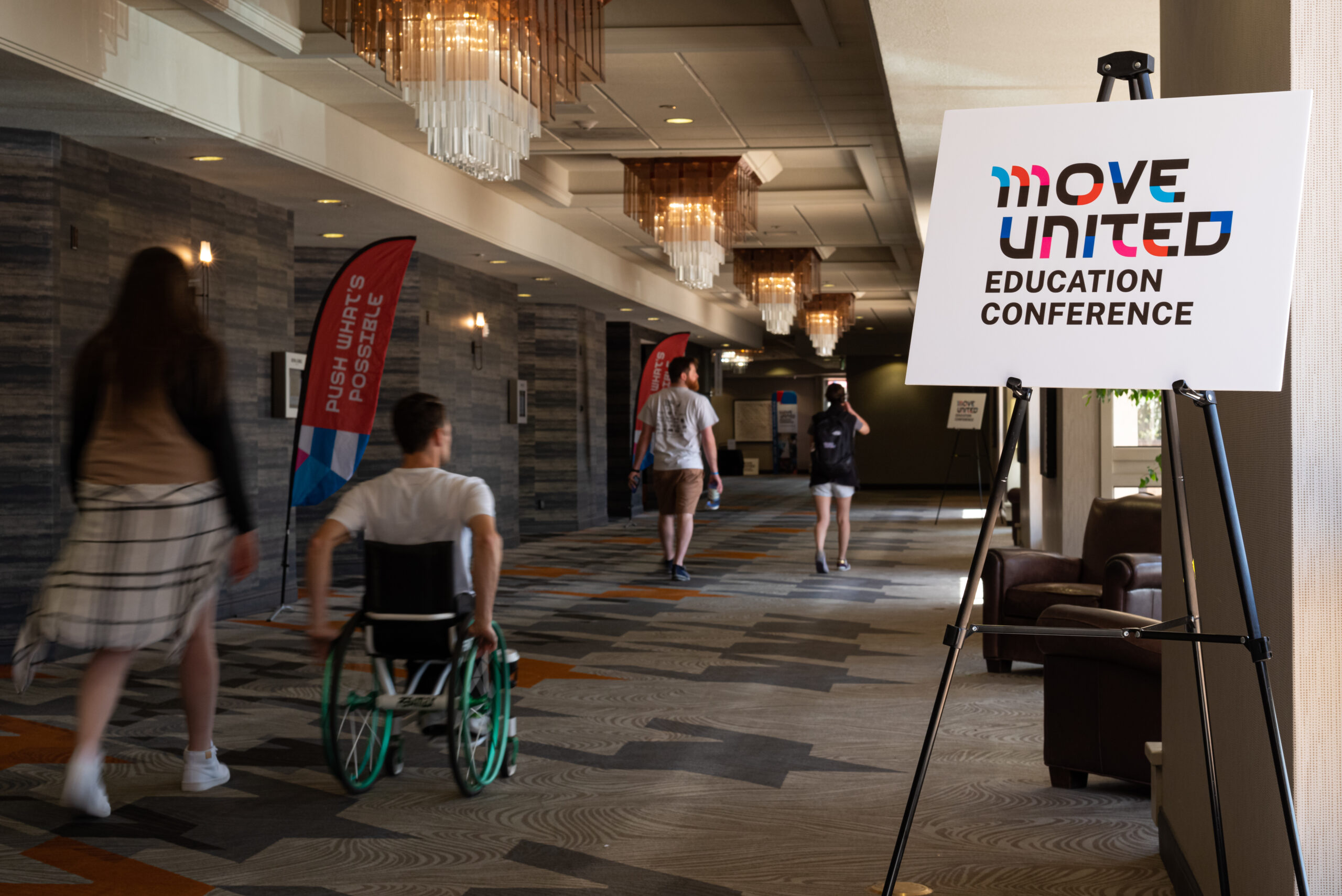 Education conference attendees rolling and walking down conference hallway, Move United Education Conference sign displayed on right of attendees