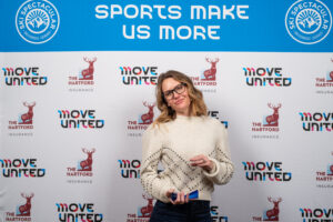 Amy Purdy holding her award in front of a banner that reads Sports Make Us More and the Move United logo