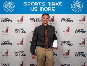 Jeff Inouye holding his award in front of a banner with text reading Sports Make Us More and Move United logo