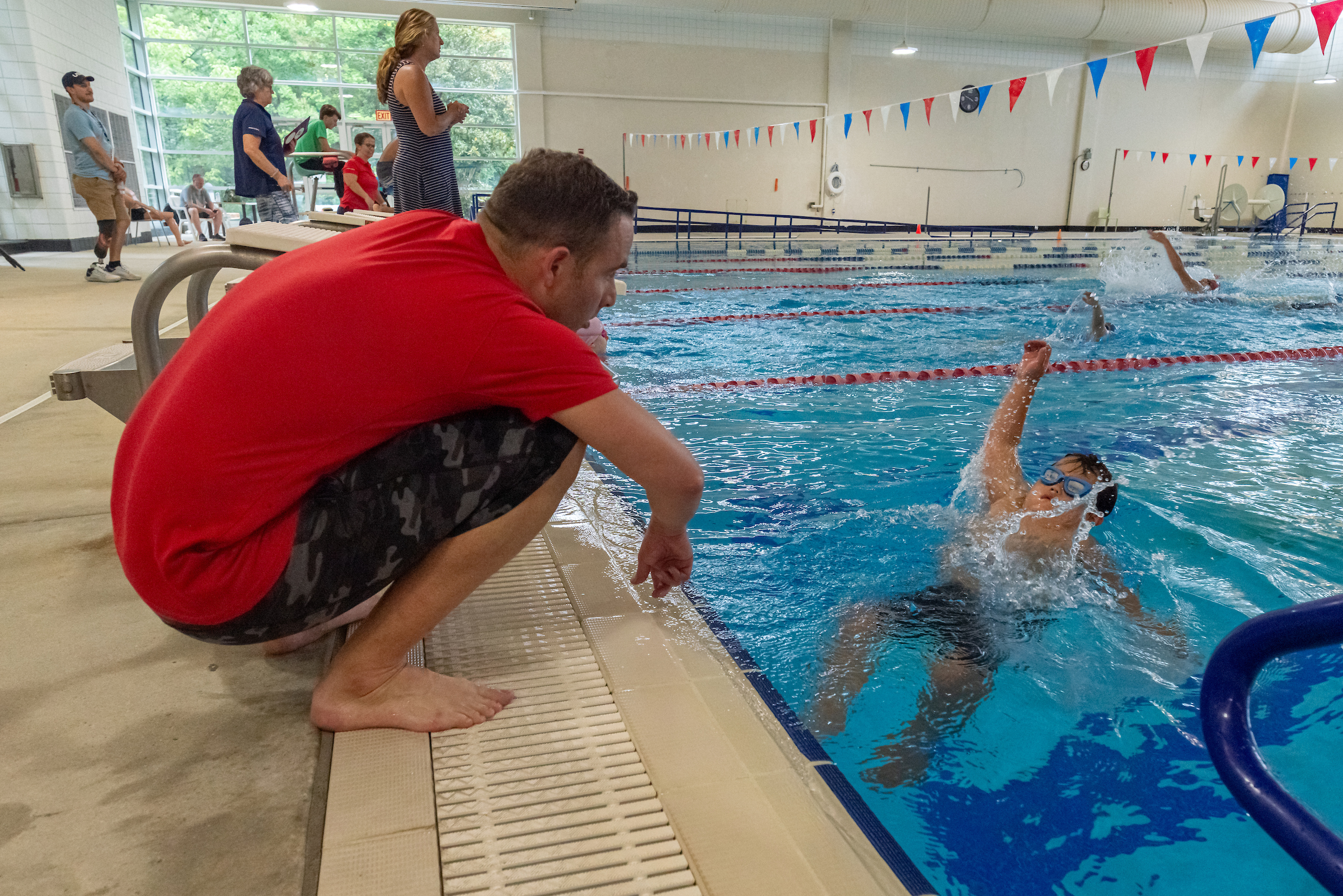 Young boy doing the backstroke while being coached by a man poolside wearing a red shirt.