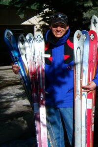 Jeff standing in a blue ski jacket, hat, and jeans, holding multiple skis on both side of himself.