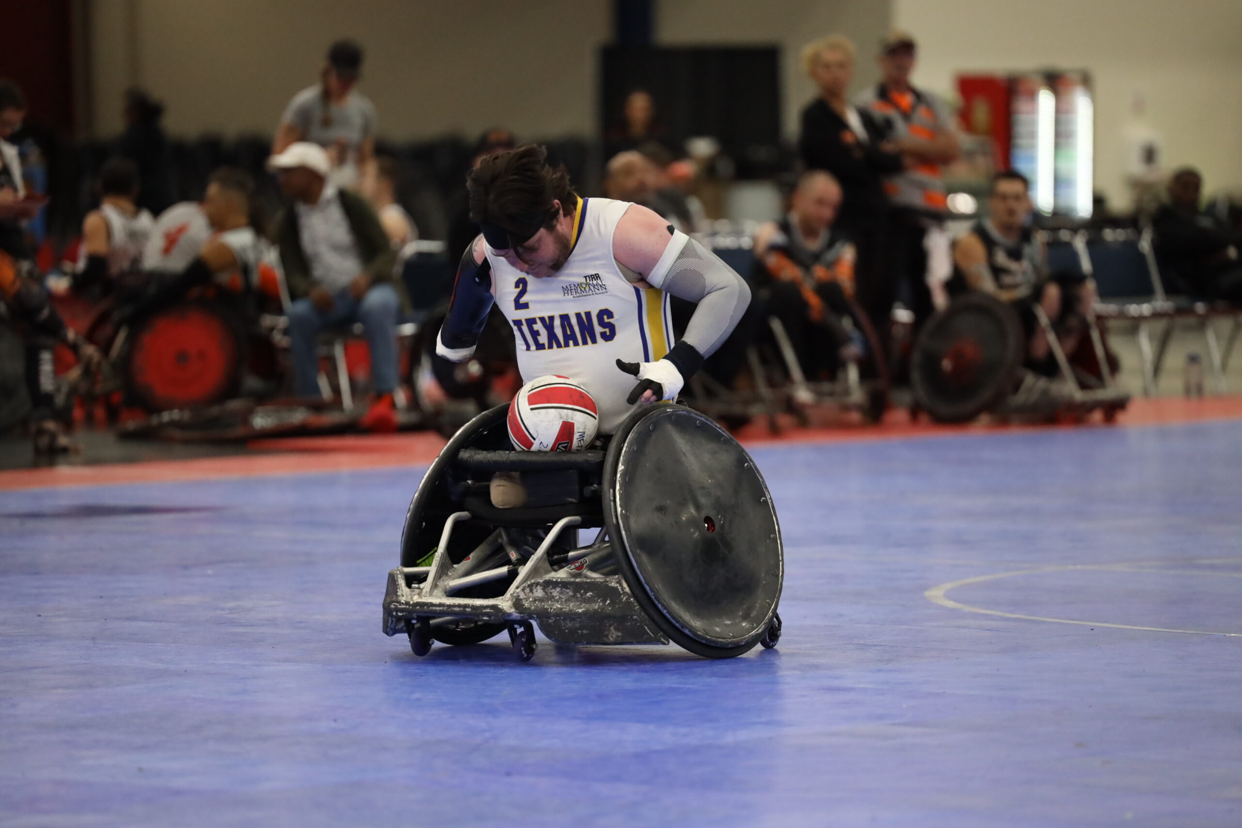 Photo of Timothy Brown pushing his rugby wheelchair during a tournament. Tim is wearing a white TIRR Texans #2 jersey.