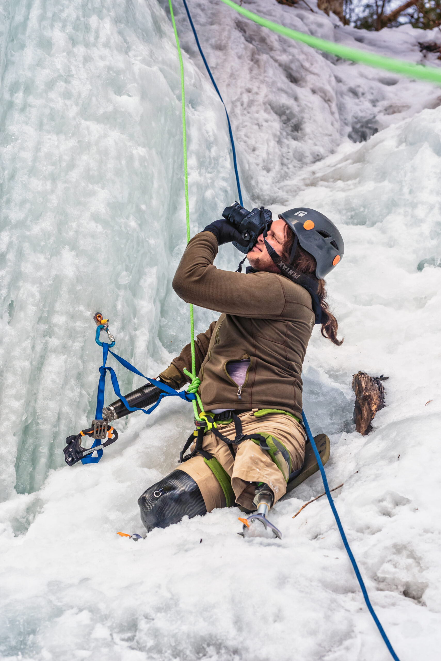 Photo of Timothy Brown sitting on an icy ledge, pointing his Pentax camera up at a fellow climber. Tim is wearing full ice climbing equipment, including three specialized ice climbing prosthetics.