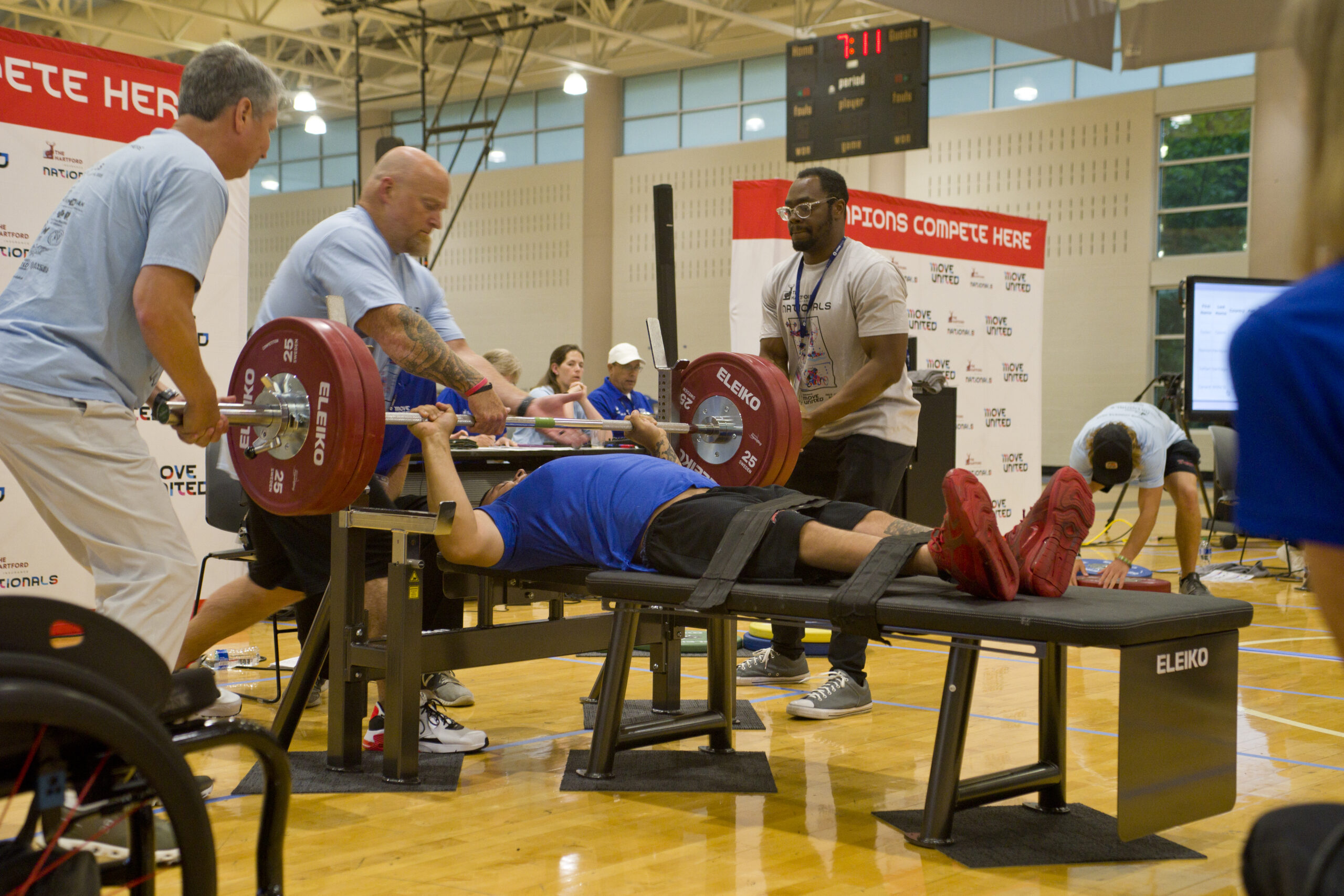 Powerlifting athlete laid down and strapped on bench. Bar in press position. Three volunteers assisting.