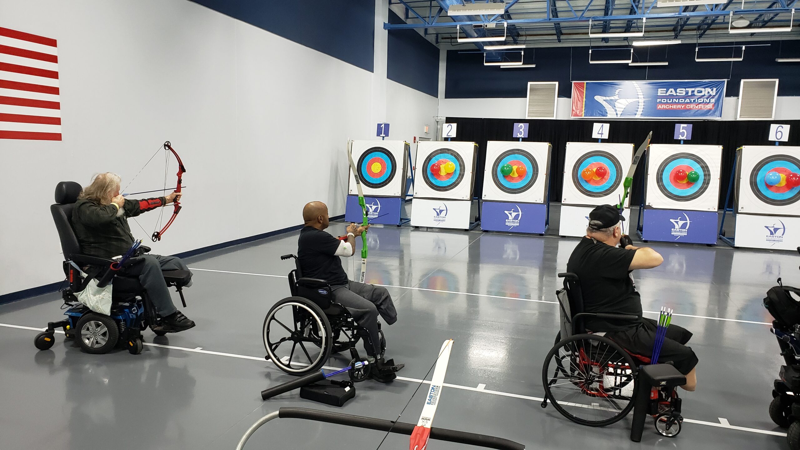 Three archers in wheelchair draw back their bows and shoot on an indoor range