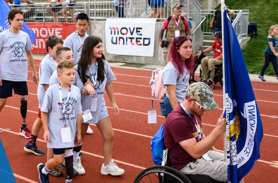 Group of adaptive athletes on a track, one in front is carrying a flag. There is a Move United banner in the background.