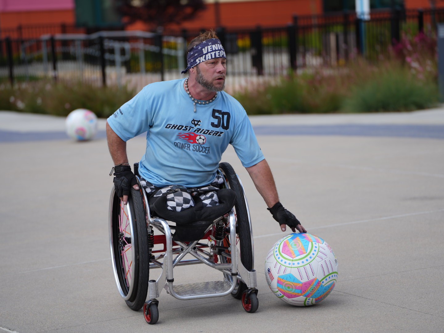 An athlete in a wheelchair, wearing a light blue shirt and bandana has their hand on a power soccer ball on the ground