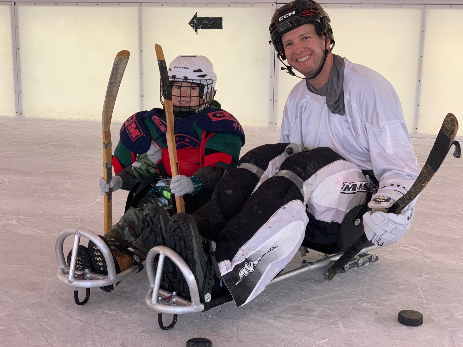 An adult and young sled hockey player pose for a picture on ice, in their gear.